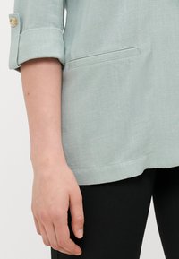 Himmelblaue, strukturierte Bluse mit hochgerollten Ärmeln und einem Knopfdetail. Verfügt über eine Tasche und wird mit schwarzen Leggings kombiniert.