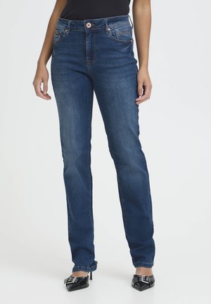 Osoba nosiaca tmavomodré džínsové nohavice s vysokým pásom a rovnými nohavicami, kombinované s čiernymi lodičkami so špicatou špičkou a čiernym topom, stojaca na bielom pozadí.