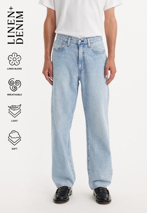 Man draagt lichtblauwe rechte jeans, wit T-shirt en zwarte schoenen, met iconen die linnenmix, ademend, licht en zachte stof aangeven.