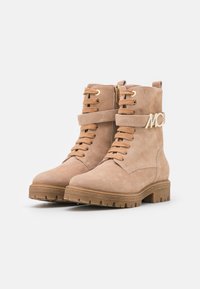 Bottines en daim beige avec des finitions en métal doré, conception à lacets, zip sur le côté et une sangle avec logo autour de la cheville. Semelle épaisse.