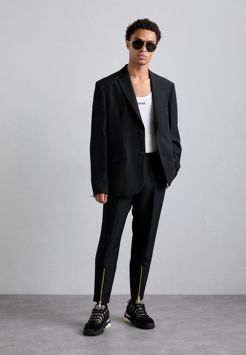 Zwarte op maat gemaakte pakken met een blazer met één knoop, gecombineerd met slim-fit broeken met gouden ritsen. Draagt een witte tanktop en zwarte sneakers.