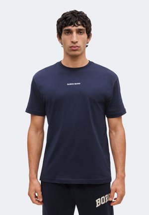 Homme aux cheveux courts et bouclés portant un t-shirt Björn Borg bleu marine et un pantalon foncé, debout devant un fond clair uni.