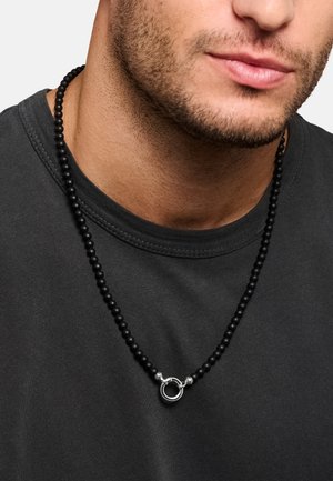 Collana nera con perline e chiusura circolare in argento indossata sopra una maglietta girocollo grigio scuro sul collo e la linea della mascella di un uomo.
