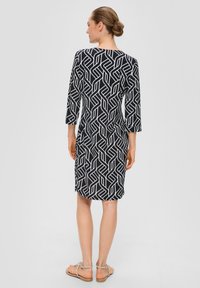 Robe à motifs noir et blanc avec des manches trois-quarts, longueur genou, présentant un design géométrique et un tissu doux. Portée avec des sandales plates.