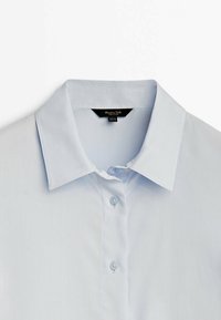 Chemise bleu clair à col avec deux boutons visibles et une étiquette de marque noire à l'intérieur de l'encolure sur fond blanc.