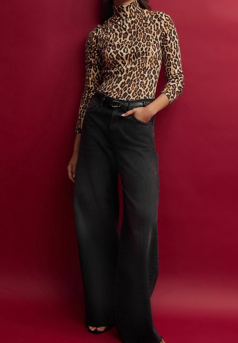 Femme portant un haut à manches longues imprimé léopard, rentré dans un pantalon noir taille haute à jambes larges avec ceinture noire, debout devant un fond rouge.