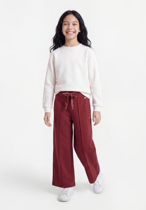Smilende pige med langt mørkt hår står iført hvid langærmet top, vidde røde bukser med snøre og hvide sneakers mod hvid baggrund.
