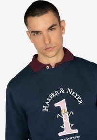Felpa blu navy con colletto bordeaux; presenta una grafica del numero “1” in rosa, vicino a un casco da cantiere giallo e mazze da golf incrociate. Testo: “HARPER & NEYER.”