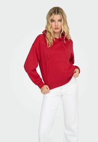 Jeune femme aux cheveux blonds ondulés portant un sweat à capuche rouge et un pantalon blanc, tenant délicatement l'ourlet du sweat avec une main.