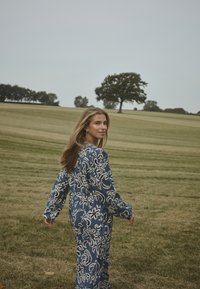 Blauer Jumpsuit mit weißem Blumenmuster, langen Ärmeln und lockerer Passform, im Freien in einer grünen Landschaft gezeigt.