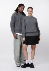 Un homme et une femme portant des pulls gris assortis, la femme en jupe plissée noire avec des mocassins, l'homme en jean bleu clair et baskets, debout côte à côte.