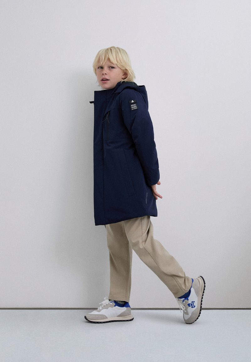 Blond kind dat loopt, gekleed in een donkerblauwe hoodie, beige broek en witte sneakers met blauwe accenten tegen een effen lichte achtergrond.