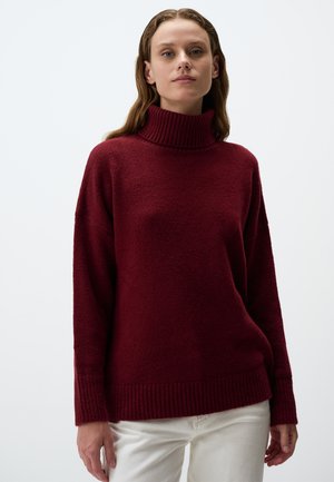 Burgunderfarbener gestrickter Rollkragenpullover mit plüschiger Textur, angeschnittenen Schultern und gerippten Bündchen und Saum; kombiniert mit weißen Hosen.