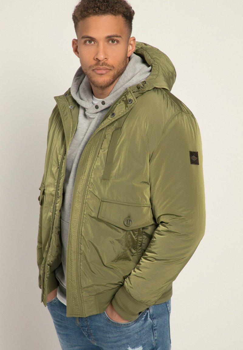 Giacca puffer verde oliva con cappuccio, dotata di chiusura con zip e bottoni, due tasche frontali e polsini a costine. Indossata sopra un felpa grigia.