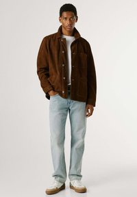 Hnedá semišová bunda s golierom, zapínaním na gombíky a prednými vreckami, v kombinácii so svetlomodrými džínsami a bielymi teniskami. Hladká textúra.