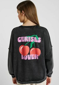 Un sweat à capuche noir surdimensionné avec un design graphique au dos, comportant du texte rose indiquant « AMATEUR DE CERISES » et deux cerises rouges avec des tiges vertes.