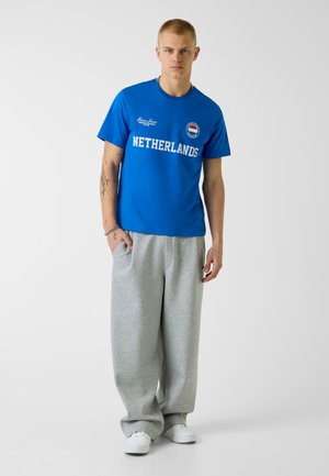 Jonge man staat met handen in de zakken, draagt een blauw T-shirt met "NETHERLANDS", grijze brede joggingbroek en witte sneakers tegen een witte achtergrond.