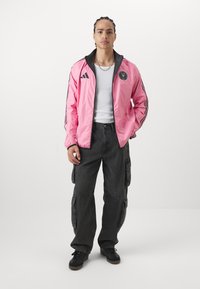 Giacca a vento rosa con accenti neri e un logo, abbinata a pantaloni cargo scuri e sneakers nere. Tessuto liscio, chiusura a zip frontale, polsini elasticizzati.
