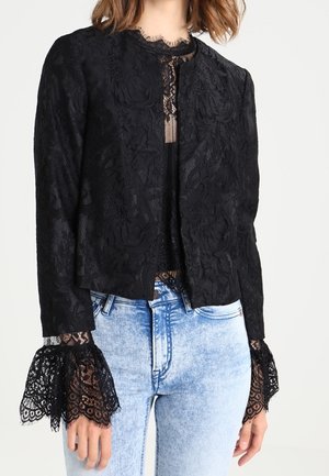 Blouse courte en dentelle noire à manches longues avec motifs floraux et poignets évasés, portée avec un jean taille haute bleu clair.