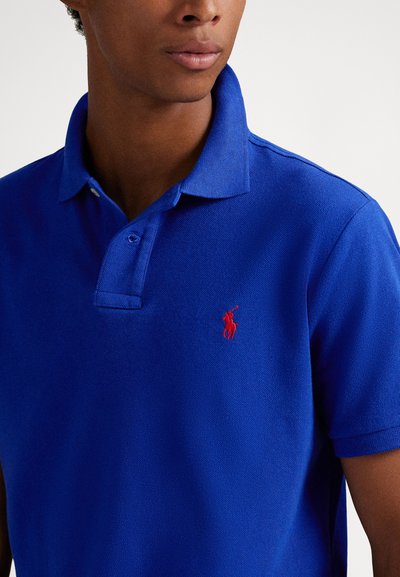 Polo azul royal feito de tecido texturizado, com colarinho clássico e um pequeno logotipo vermelho bordado no peito esquerdo.