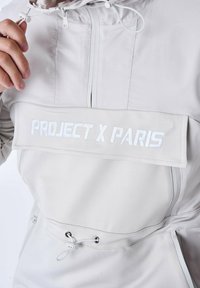 Lätt, krämfärgad jacka med dragsko i huvan, framlogo-tryck i vitt och kontrasterande dragkedjor. Har en strukturerad tyg.