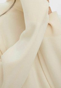 Gros plan sur les manches d'un pull tricoté couleur crème avec des poignets côtelés et un détail de couture visible près de l'épaule.