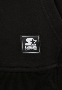 Černá mikina s kapucí, která má textilní nášivku s logem "Starter Black Label", kontrastní bílý text a design na texturovaném pozadí.