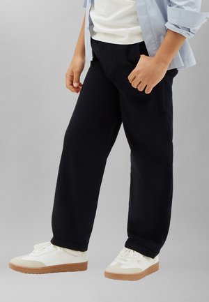 Persona che indossa pantaloni neri a gamba larga, sneakers bianche con suola di gomma, camicia bianca e camicia a maniche lunghe azzurro chiaro con polsini risvoltati.