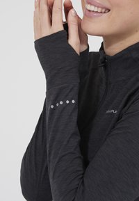 Frau lächelt, die Hände nahe am Gesicht gehalten, trägt ein dunkles, langärmeliges Zip-up-Oberteil mit Daumenlöchern und reflektierenden sechseckigen Details an den Ärmeln.