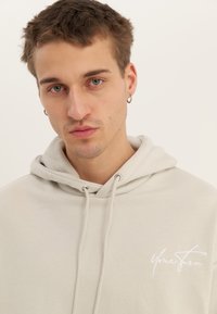 Mlad moški s kratkimi rjavimi lasmi in zelenimi očmi, oblečen v svetlo bež pulover s kapuco z napisom "Your Turn" na prsih, nevtralna ozadje.