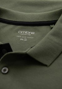 Olivgrön polo skjorta i 100% bomull, med svart krage och en etikett som visar varumärke och storleksinformation.