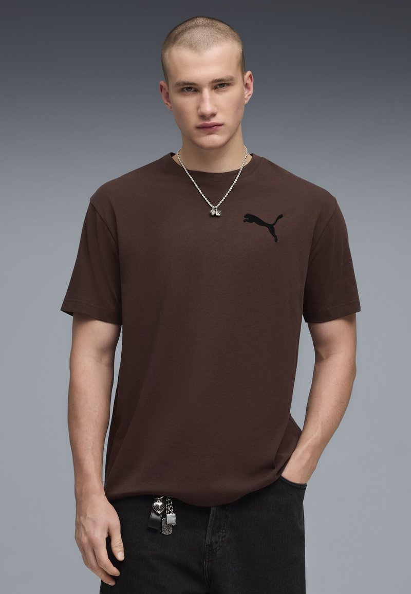 Jeune homme avec une coupe rasée, portant un t-shirt marron Puma, un collier argenté et un pantalon noir avec des accessoires porte-clés, debout devant un fond gris.