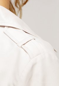 Jasno beżowy strój z wyraźnym kołnierzem, teksturowanym materiałem oraz charakterystycznymi detalami w postaci pasków na ramionach, ukazujący elegancki, minimalistyczny design.