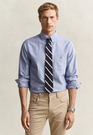 Homme portant une chemise bleu clair boutonnée avec manches retroussées, une cravate rayée bleu marine, un pantalon beige et une montre-bracelet argentée sur fond uni.
