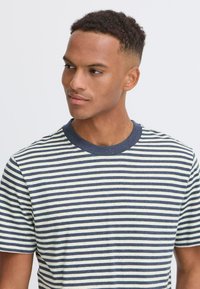 Dryžuotas marškinėlis, kuriame yra tamsiai mėlynų ir pieniškų horizontalių dryžių, apvalus iškirpimas ir trumpi rankovės. Audinys atrodo minkštas su tekstūruotu paviršiumi.