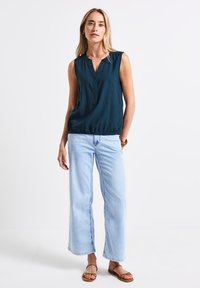 Blouse sans manches vert foncé avec un décolleté en V et un ourlet élastique, associée à un jean à jambes larges bleu clair et à des sandales à lanières marron.