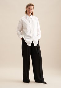 Witte button-up blouse met een puntkraag en lange mouwen, gecombineerd met zwarte wijde broek en zwarte puntige schoenen.