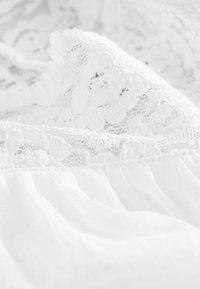 Tissu en dentelle blanche avec des motifs floraux cousus sur un tissu blanc doux et fluide, mettant en valeur la texture et les détails délicats.