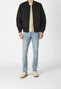 Chaqueta bomber negra con textura suave, cierre de cremallera y puños acanalados, combinada con jeans azul claro y sneakers beige.