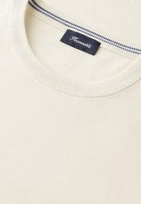 Cuello de suéter de punto color crema con etiqueta de la marca Pañcomoble en azul marino y sutiles rayas azules en el interior del cuello.