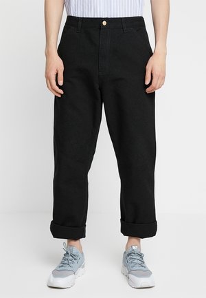 Person iført sorte bukser med opsmøg, lyseblå og hvide sneakers og en skjorte med striber, der er stukket ned, stående mod en ensfarvet baggrund.