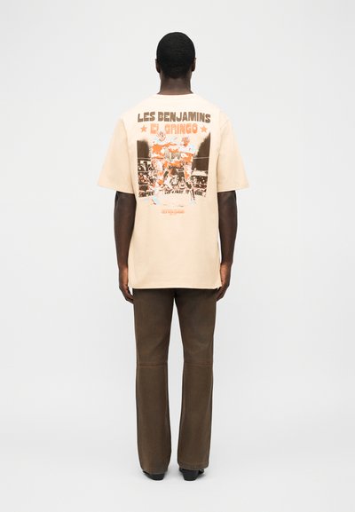 Άνδρας που φοράει μία μπεζ υπερμεγέθη μπλούζα με γραφικό πυγμαχίας και το κείμενο "Les Benjamins El Gringo" στην πλάτη, σε συνδυασμό με καφέ παντελόνι και μαύρα παπούτσια.