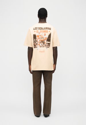 Muž v béžovom nadrozmernom tričku s boxerskou grafikou a textom "Les Benjamins El Gringo" na chrbte, v kombinácii s hnedými nohavicami a čiernymi topánkami.