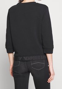 Černá mikina s kulatým výstřihem, tříčtvrtečními rukávy a strukturovaným pasem s logem. Kombinována s tmavými džínami.