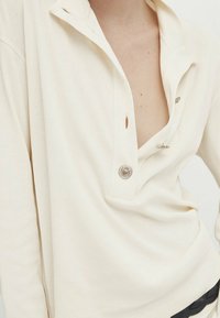 Persona che indossa una camicia a maniche lunghe di un bianco sporco con i bottoni parzialmente sbottonati, che rivelano il petto superiore e la clavicola.