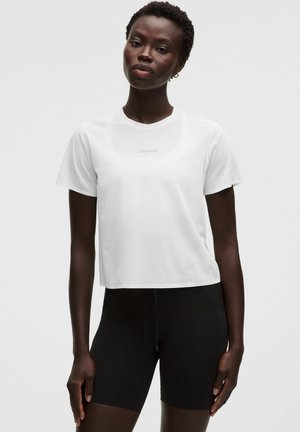 Vrouw draagt een effen wit t-shirt met korte mouwen en zwarte nauwsluitende shorts, staand tegen een lichte achtergrond.