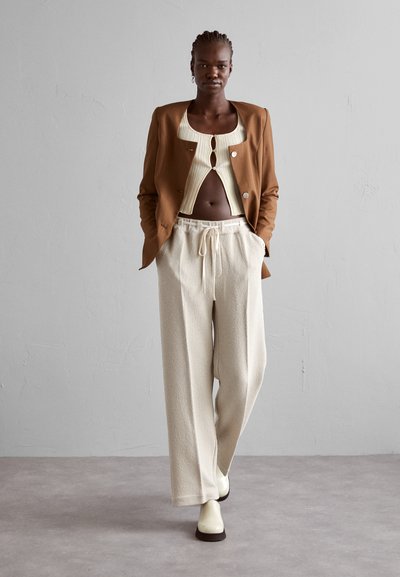 Γυναίκα με πλεξούδες φοράει ένα καφέ σακάκι, κρεμ κρουαζέ μπλουζάκι, φαρδιά μπεζ παντελόνια και ανοιχτόχρωμα μποτάκια, περπατώντας σε γκρίζο δάπεδο.