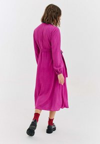 Różowa sukienka z długim rękawem, paskiem w talii i wzorem w groszki. Luźny materiał sięga do połowy łydki, zestawiona z czarnymi butami i czerwonymi skarpetkami.