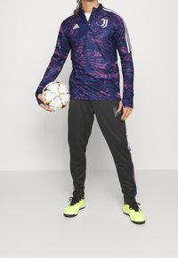 Liila kuvioitu pitkähihainen paita, jossa valkoiset raidat ja logo, yhdistetty mustiin housuihin, joissa on värikkäät sivukoristeet, ja kirkkaan keltaiset nappulakengät.