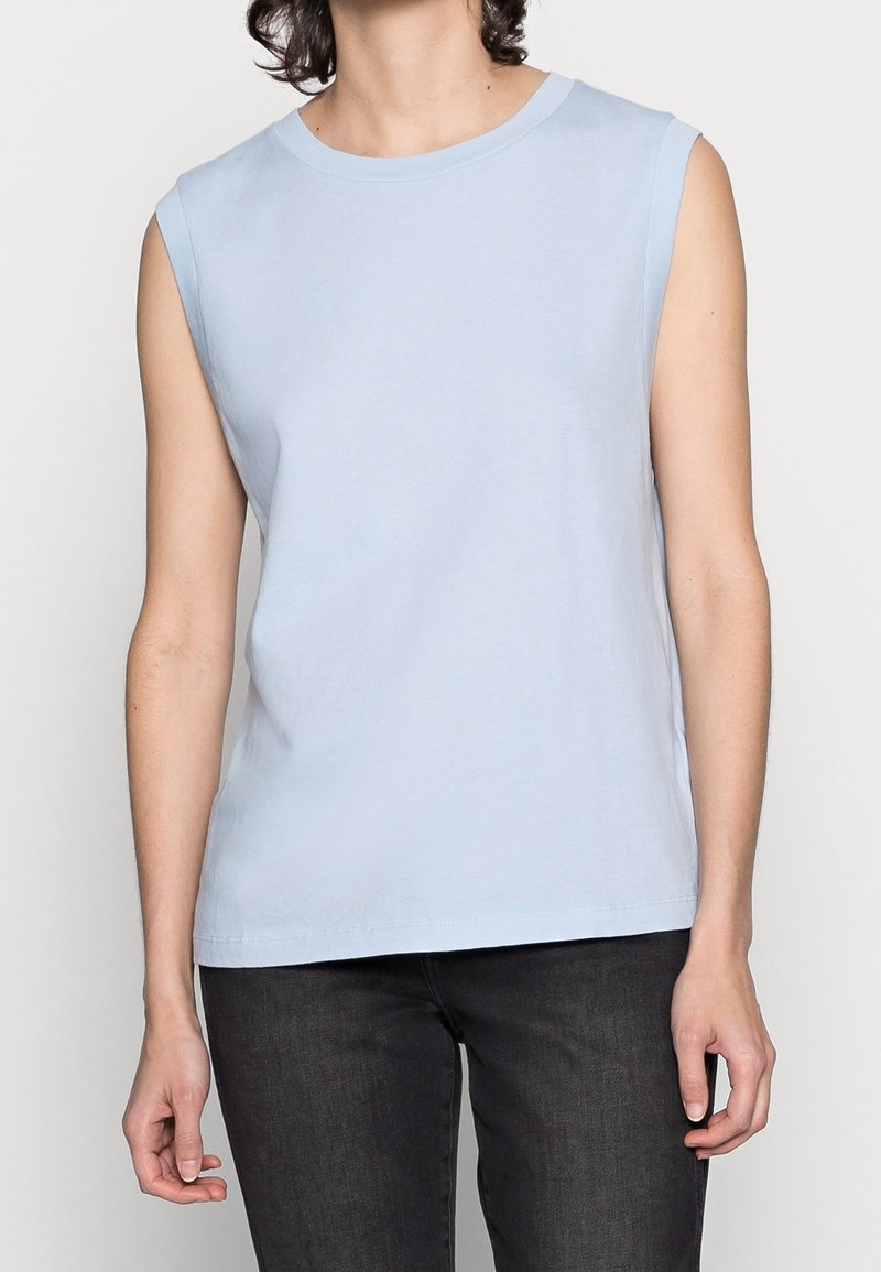 Personne portant un haut sans manches bleu clair et un jean noir, debout les bras détendus le long du corps devant un fond uni.
